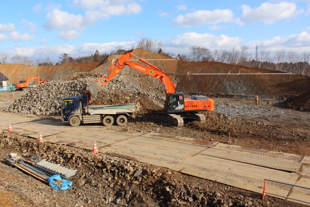 広い工事現場でオレンジ色のショベルカーがダンプトラックに土砂を積み込み、その周囲には作業員や重機、敷き詰められた鉄板などが見られる写真