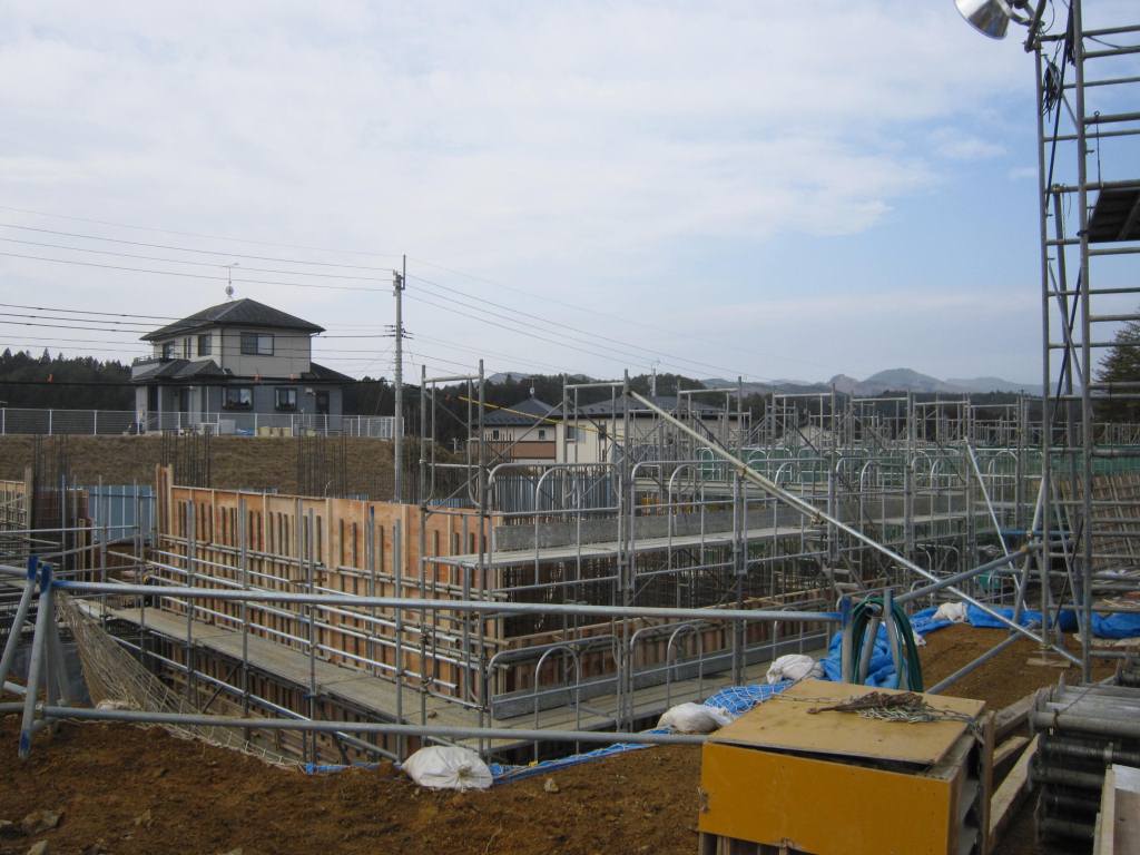 鉄パイプで組まれた足場と型枠が整然と並び、住宅と山並みが背景に見える建築中の構造物の写真