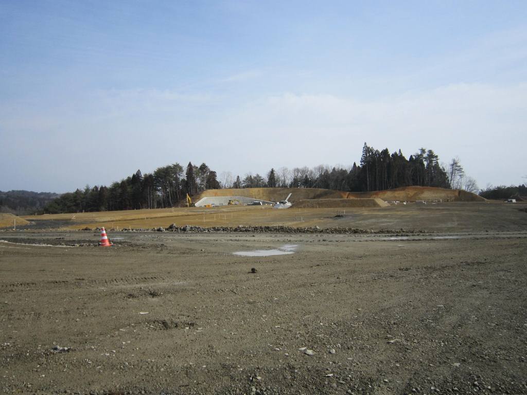 広い空き地の奥に建設中の工事現場があり、その周囲に木々が立ち並んでいる様子を写した写真
