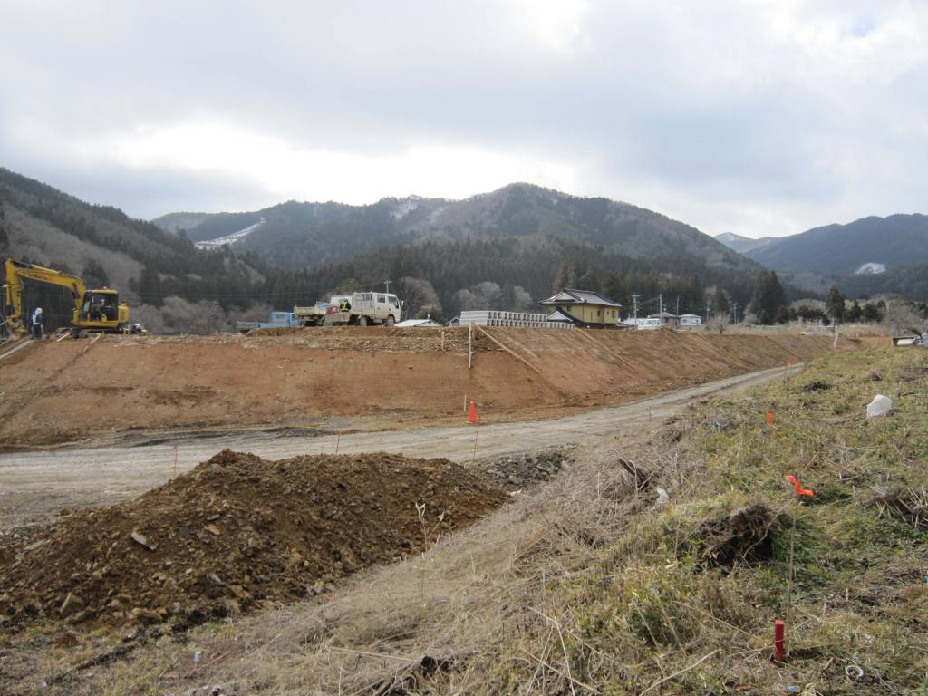 手前に草地と未舗装の道路、その奥に土を盛って整地された高台が広がり、高台の上にはショベルカーやトラックなどの工事用車両がある造成工事現場の写真