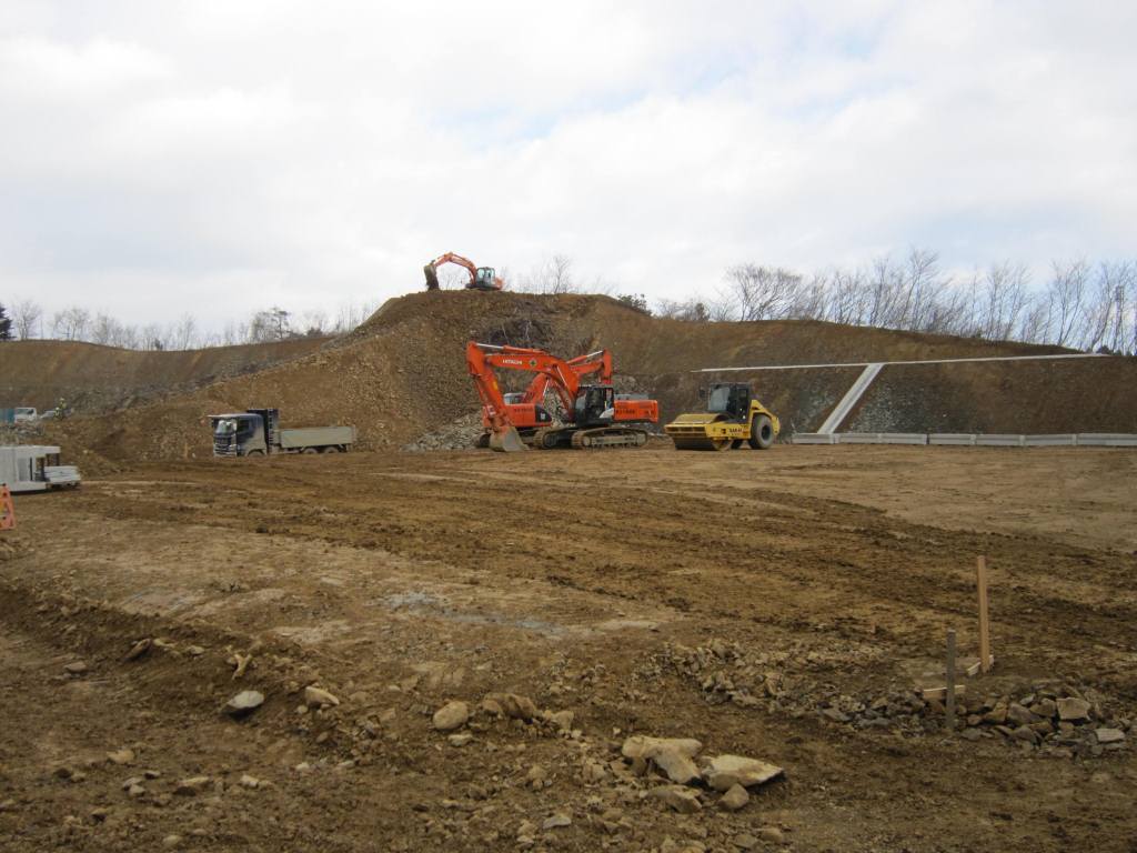ショベルカーやダンプトラックが稼働し、土を掘削・運搬・整地している広大な造成地で、法面にはコンクリート擁壁が設けられている工事現場の様子を捉えた写真