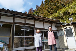 奥に山が見え、工房の前で厨さんと及川美樹さんがエプロンをして並んでいる写真