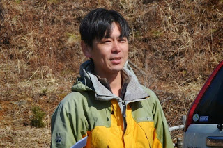 枯草が覆う斜面の前で話をしている、黄色と緑の上着を着た鈴木卓也さんの写真