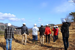 白いヘルメットをかぶった人達が、枯れ草の広がる山に集まっている写真