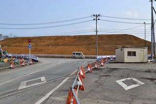 中央には交差点があり、道路が左右に分かれ、交差点の手前と奥にはオレンジ色のコーンが並べられ、左側には「止まれ」の標識、右側には小さなプレハブの建物が見える道路工事の写真