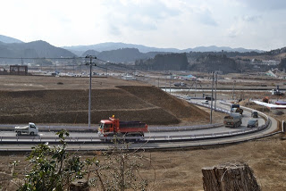 手前には曲がった道路が通ってトラックや乗用車など複数の車両が走行し、道路沿いには電柱が並んでいる写真