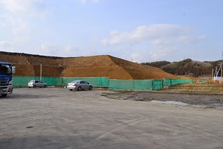 大きな土の山が緑色のネットで覆われ、左側に青いトラック、中央に白い車、右側にももう1台の白い車が停まっている建設現場の写真