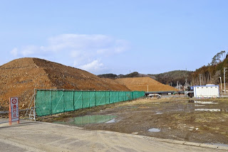 大きな土や砂利の山が緑色のフェンスで囲われ、地面はぬかるんでいて、水たまりが点在している建設現場の写真