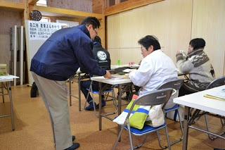 参加者の方が座っている机に関係者の方が近づいて説明をしている写真