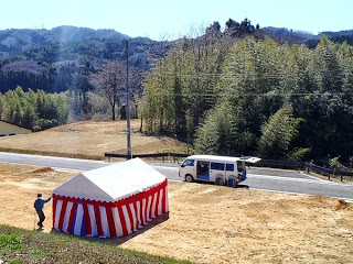 整備された土地に白と赤のテントが設営され、近くに作業員と白いバンが見える行事の準備中の写真