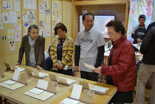 4人の人がテーブルの周りに立って、テーブルの上の作品を手に取ってみたり、話しをしている様子を写した写真