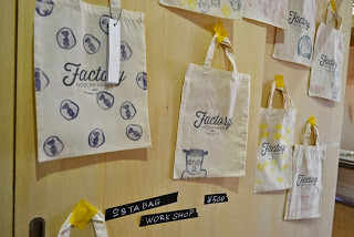 「Factory」のロゴとオリジナル絵やスタンプが押されている白い布製のエコバッグが壁に複数枚掛けられ展示されている写真