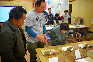 机に展示されている作品を覗き込みながらみている2名の人とスタッフの男性の写真