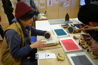 机の上に色々なインクとローラーが置かれていて、男性がインクを使ってスタンプを使い作品を作っている