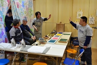 L字型の長机にスタンプ台とスタンプアイロンが置かれ、奥の男性2名が手を前に出しながらタッチのようなしぐさをしており、隣にスタッフの男性2名が座っている写真