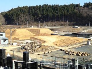 山に囲まれた平地に高さの異なる土の山が3つ並んでおり、整地された土が広がっている写真