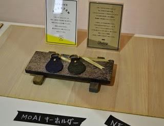 木の台に2つのMOAIキーホルダーが並べられており、奥には説明文の紙が立てられている写真