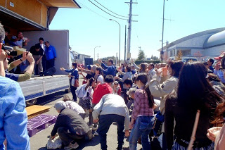 数名がトラックの荷台の舞台に立ち、餅まきを行い、集まったたくさんの人々が、地面にかがんで拾ったり、手を差し出してつかみ取ろうとしている様子の写真
