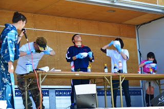 ステージ上のテーブルの後ろに4人の男女が並んで早食いチャレンジしており、その横で浴衣を着た女性がマイクを持って立っている様子の写真