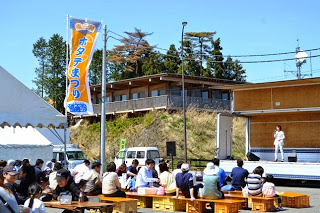 「ホタテまつり」と書かれたのぼりが立った屋外スペースで、ステージ前のベンチにたくさんの観客が座り、ステージの上の出演者を見ている写真