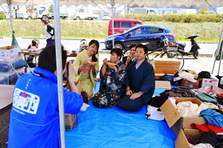 テントの下に敷かれたブルーシートの上で、4人の浴衣姿の男女が座っているところをカメラで撮影している、白い文字の入った青いジャンパーを着た男性の後ろ姿の写真