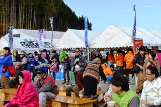 屋外に白いテントが並び、多くの来場者などで混雑したイベント会場で、木製のベンチが置かれたスペースの左端に青いジャンバーを着たNaikiさんが写りこんでいる写真