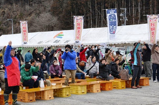 のぼりが立ち並んだ白いテントの前に設置されたオレンジ色のコンテナでつくったベンチ周辺にたくさんの人が集まり、座ったり立ったりしながら挙げていている中央あたりに、青いジャンバーを着てカメラを構えて立っているNaikiさんが写っている写真