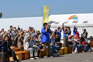 屋外でオレンジのコンテナで作ったベンチに座った人々やその周りに集まった多くの人々が拳を挙げており、中央に青いジャンバーを着てカメラを構えて立っているNaikiさんが写っている写真