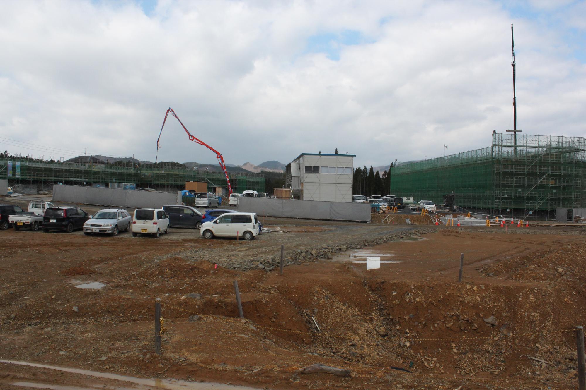 広い工事現場に多くの車両が停まり、中央に仮設建物、奥には緑の足場に覆われた建築中の建物とコンクリートポンプ車が見える工事現場の写真