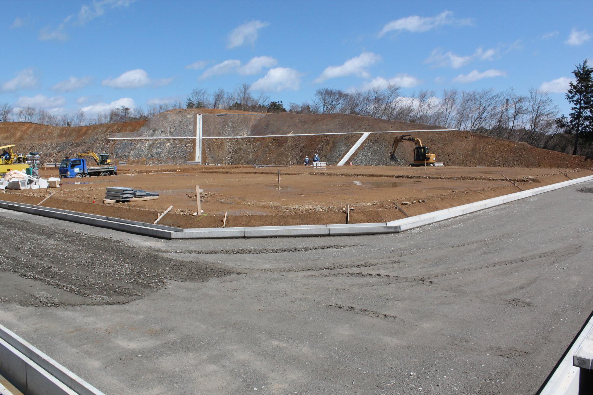 法面にコンクリートの擁壁が整備され、平坦に整地された敷地内で作業員や建設機械、資材が配置され、舗装の準備が進められている造成工事現場の様子を捉えた写真