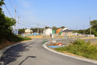 右に向かってカーブする道の先に新しい家がいくつか建設されており、資材が置かれている造成中の住宅地の写真