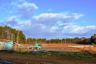 奥にはまばらな林が広がり、整地された広い土地に緑色のトラックが停まっている開発現場の写真