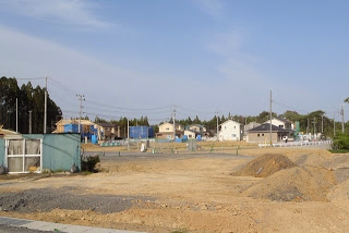 更地になった敷地内に幾つもの土の山があり、その先の造成地には完成した家や建設中の住宅が建ち並んでいる写真
