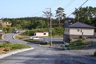 舗装前の砂利道があり、仮設の建物や新築の家、車両が見える住宅開発区域の写真
