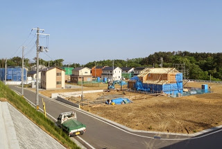 舗装された道路の奥に複数の新築住宅が建ち並び、中央には木造の建物が骨組みの状態で建設中で、周囲にはブルーシートや工事用資材が置かれている郊外の住宅地開発現場の写真