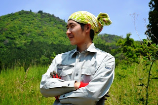 緑豊かな山と草地を背景に、頭にタオルを巻いた大岡久則さんが腕を組んで微笑んで立っている写真