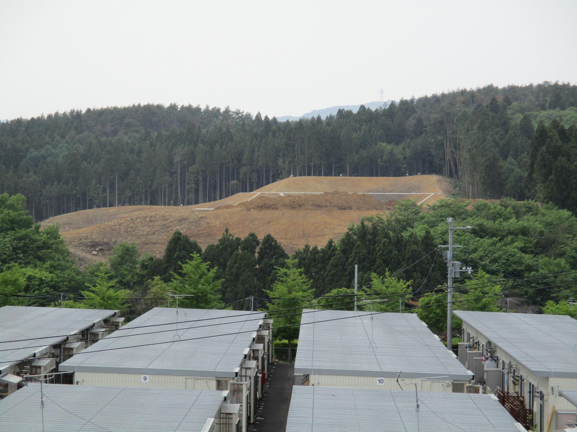 森林に囲まれた地域で、手前に規則正しく並ぶ仮設住宅群があり、その背後では防災目的の高台移転のために山を削り整地された造成地が広がり、まだ土の地肌が見える造成中の写真