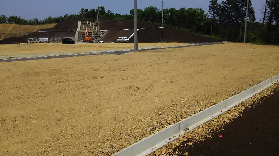 広く整地された造成地の奥で、重機と作業車が斜面の擁壁工事を行っており、周囲には舗装された道路が写っている写真