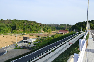 小高い山に囲まれ、左側には田畑や住宅が見え、坂を下る道路の右側に造成地が広がっている写真
