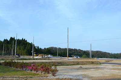花壇のツツジが咲く手前に、整地された区画と電柱が立ち並ぶ新しい住宅予定地の写真