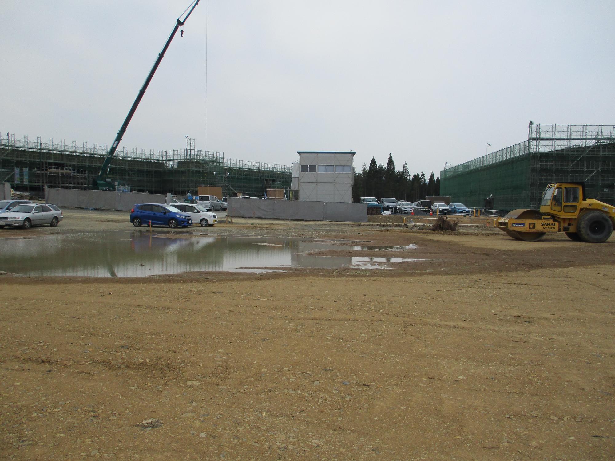 雨があがった直後の建設現場に雨水が溜まった水たまりが広がり、仮設建物と足場の組まれた建物が並んだ工事現場で重機が稼働している様子の写真