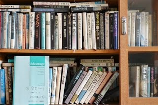 ガラス扉の書棚に古書や文学作品がぎっしりと詰まっており、手前には青い表紙のノートが立てかけられている書架の写真