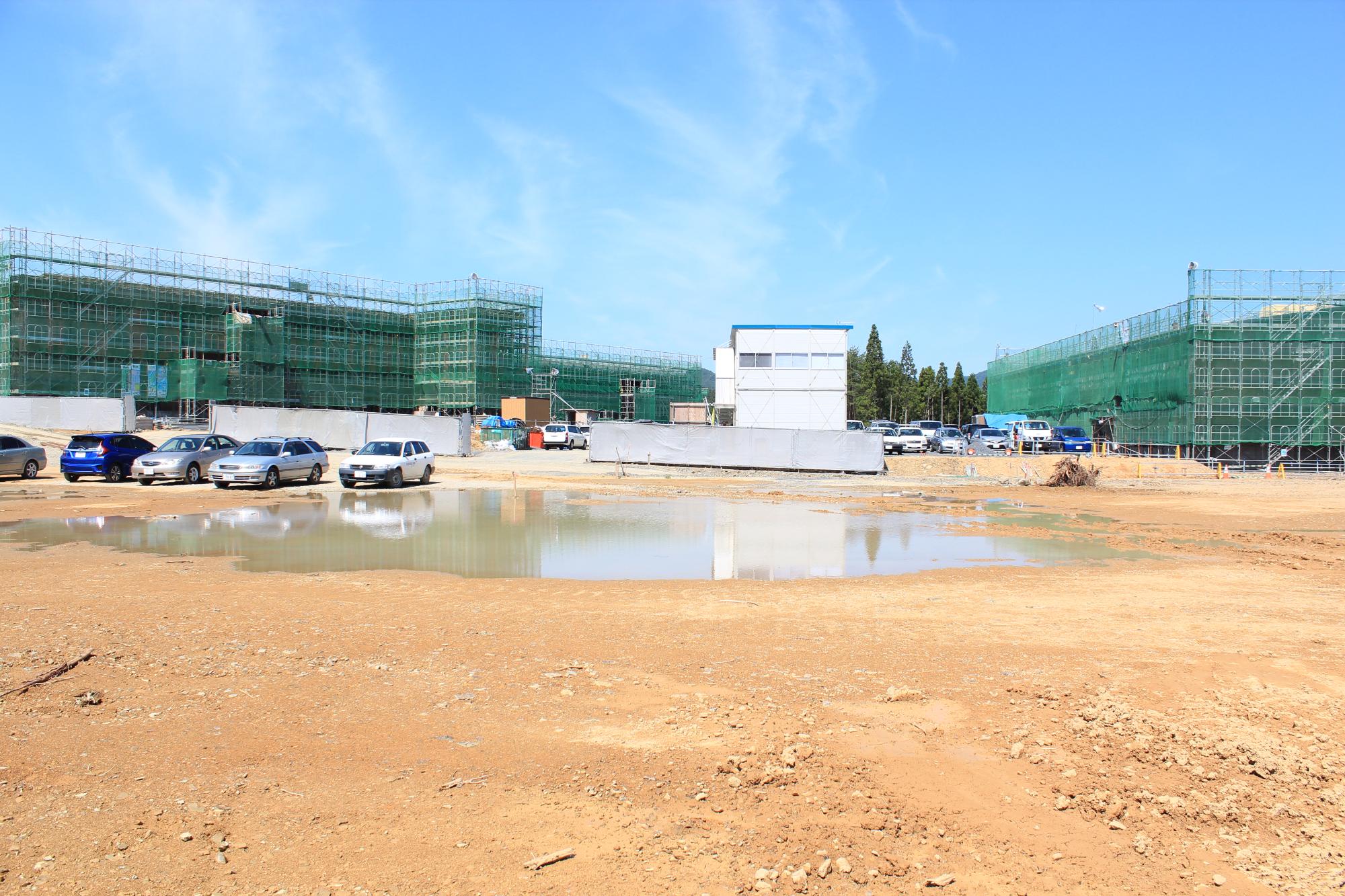 晴天の下、建設中の建物の前に複数の車が停まり、水たまりに青空が映っている建設現場の写真
