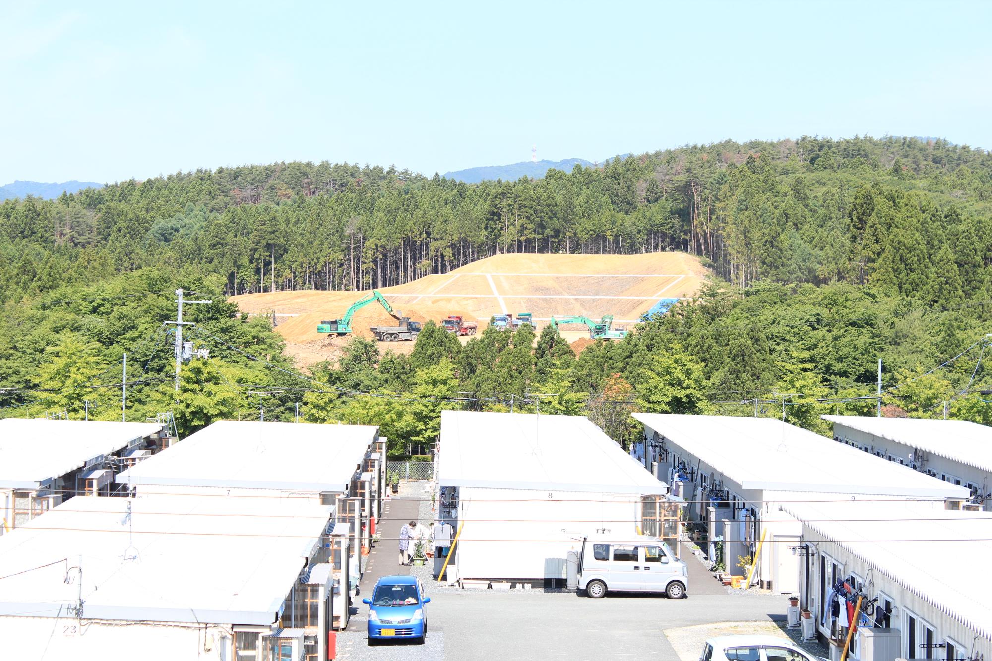 濃い緑の木々に囲まれた造成地で、多数の重機とトラックが稼働する初夏の快晴の写真
