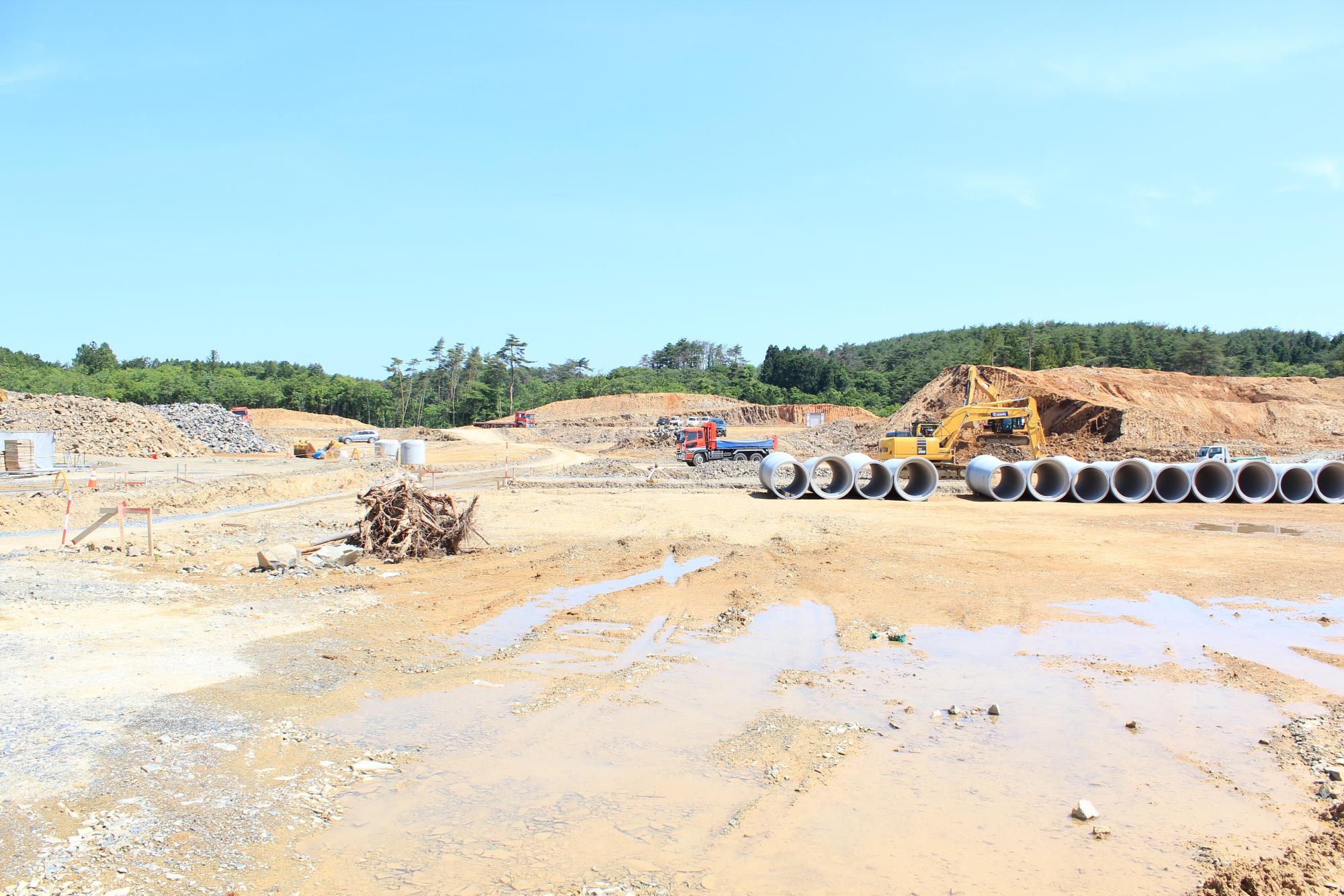 雲一つない空の下、土管が整然と並び重機とトラックが稼働している造成現場の写真
