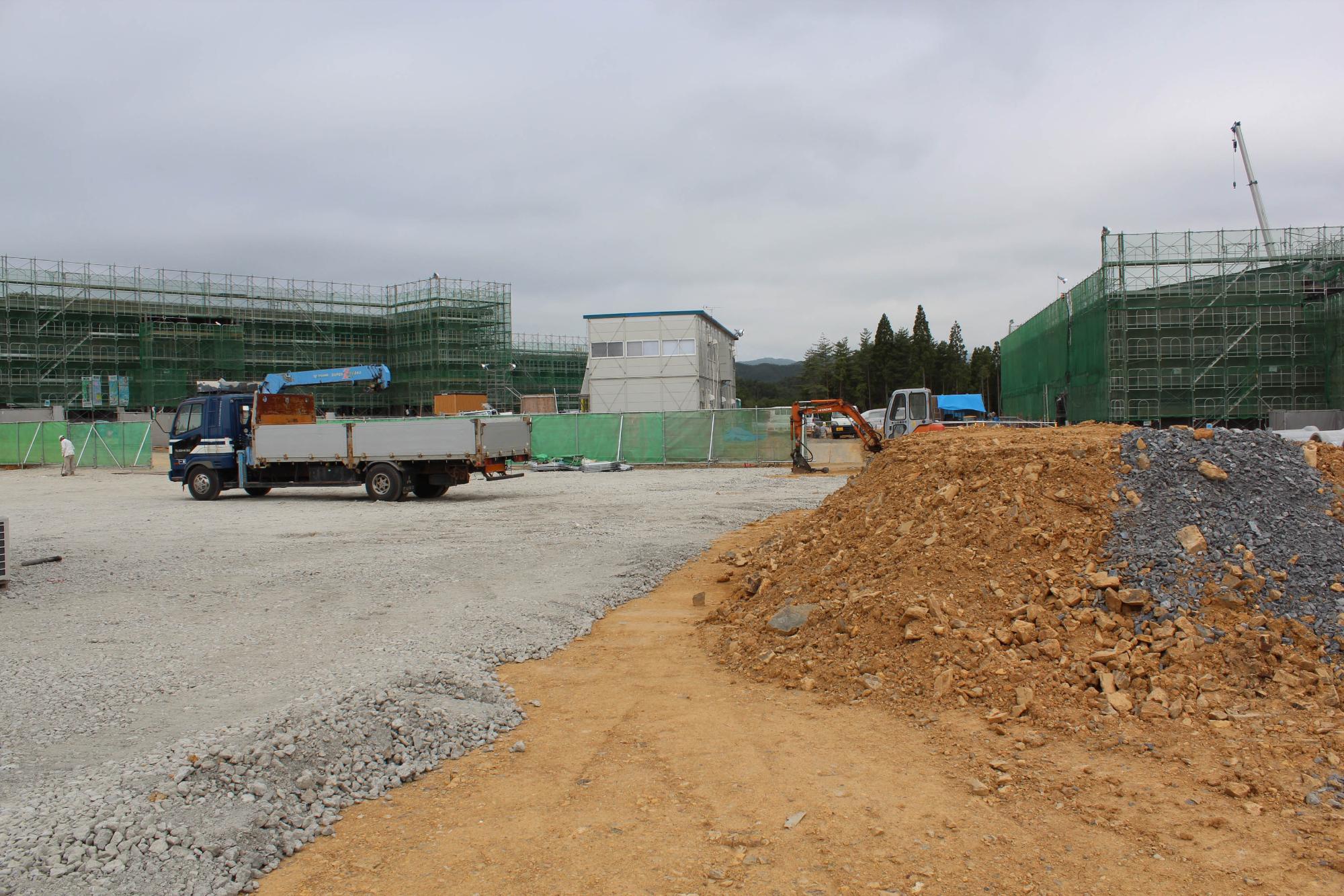 茶色い土と砕石が積まれた工事現場の手前にダンプトラックとショベルカーがあり、奥には緑の足場で囲まれた建設中の建物と仮設の白い建物が見える建設中の施設の写真