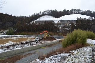雪が積もった丘を背景に、川のそばでオレンジ色のアームを持つショベルカーが土を掘り、白いトラックが停まっている工事現場の写真