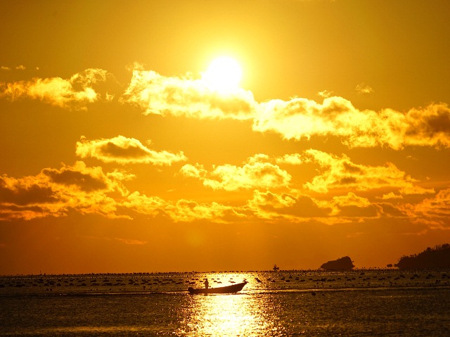 黄金色に染まった空と雲の下、太陽の光が海面に反射して輝き、1艘の小さな漁船が静かに浮かんでいる写真