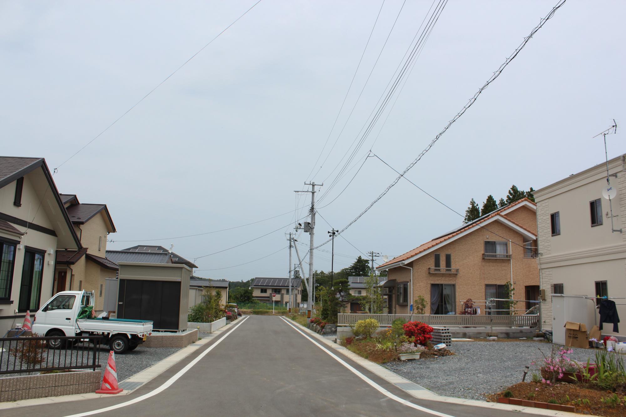 舗装された新しい住宅街の道路の両側に整った新築住宅が建ち並び、植え込みや軽トラックが見られる静かな住宅地の写真