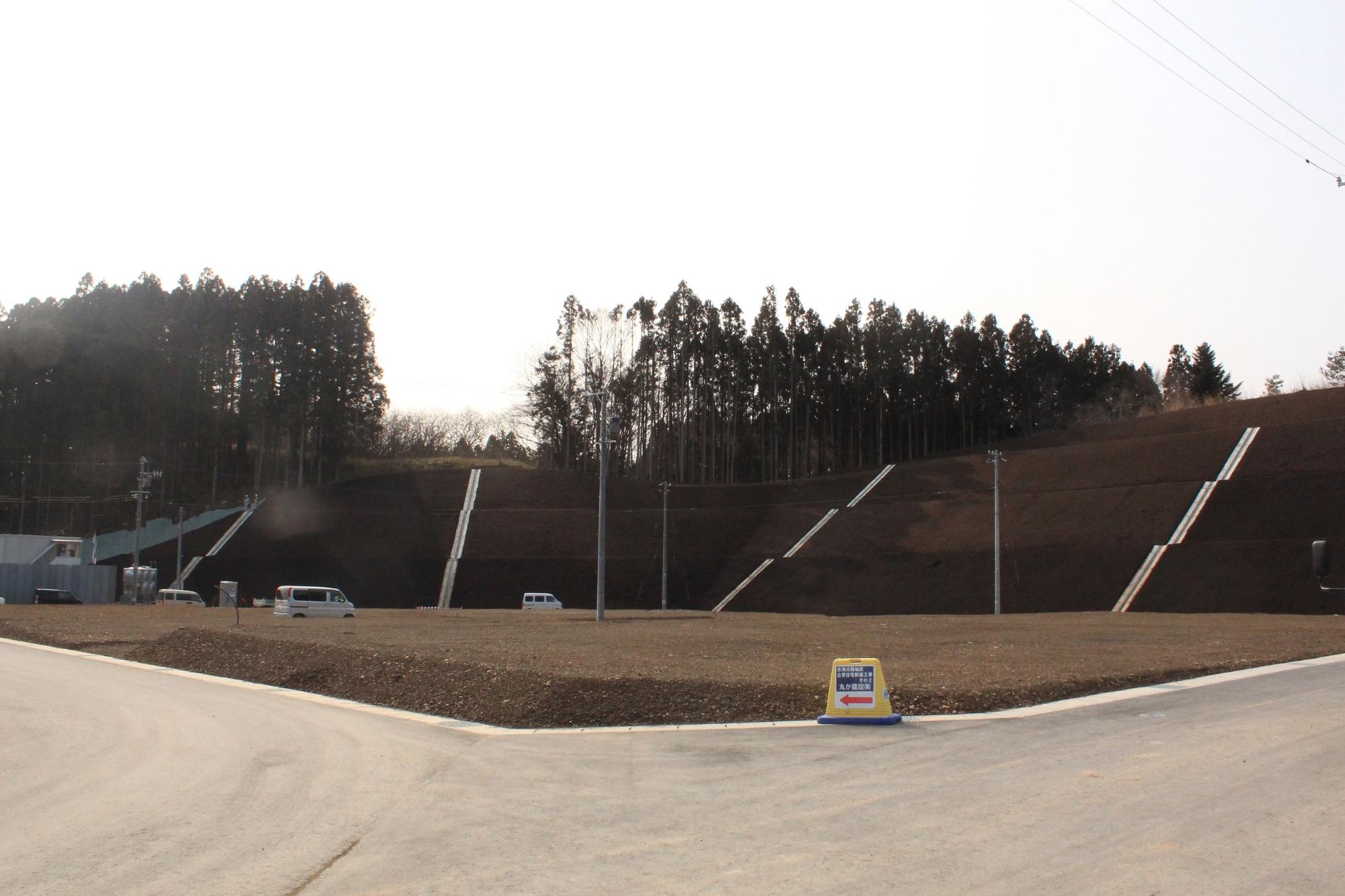 傾斜地に排水溝が設置された土盛りの法面と、その前に整備された更地と車が停まっている工事完了後の敷地の写真